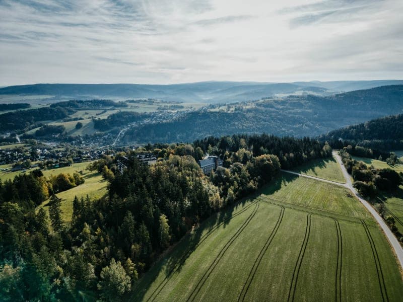 Luftaufnahme des Hotels Hoher Hahn umgeben von grünen Wiesen und Thüringer Waldlandschaft mit Hügeln im Hintergrund